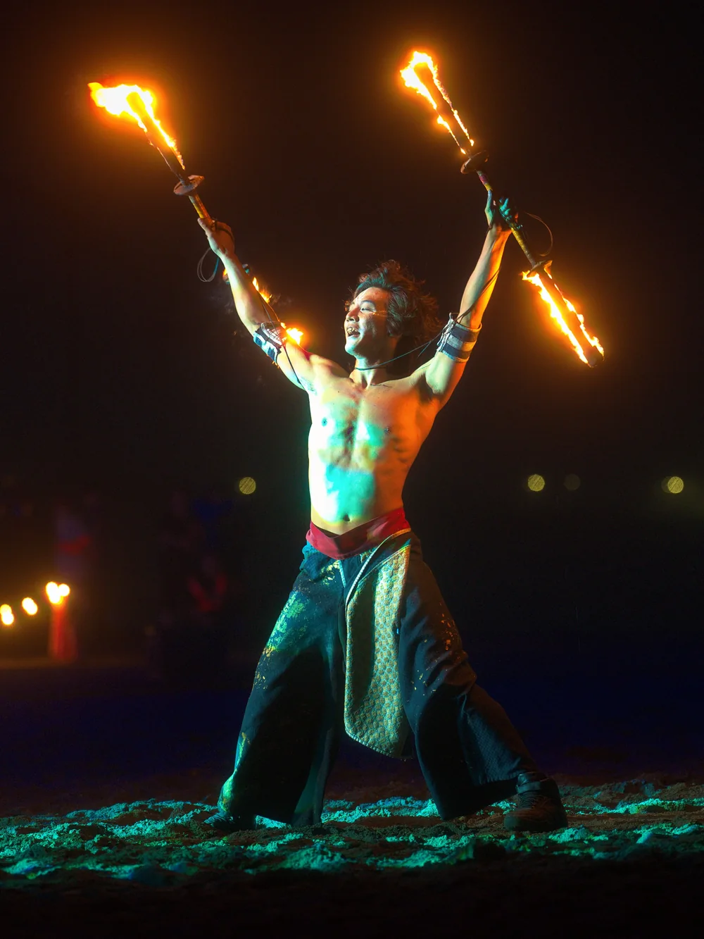 A fire performer on a dark beach holds two flaming torches aloft, string lights glowing behind