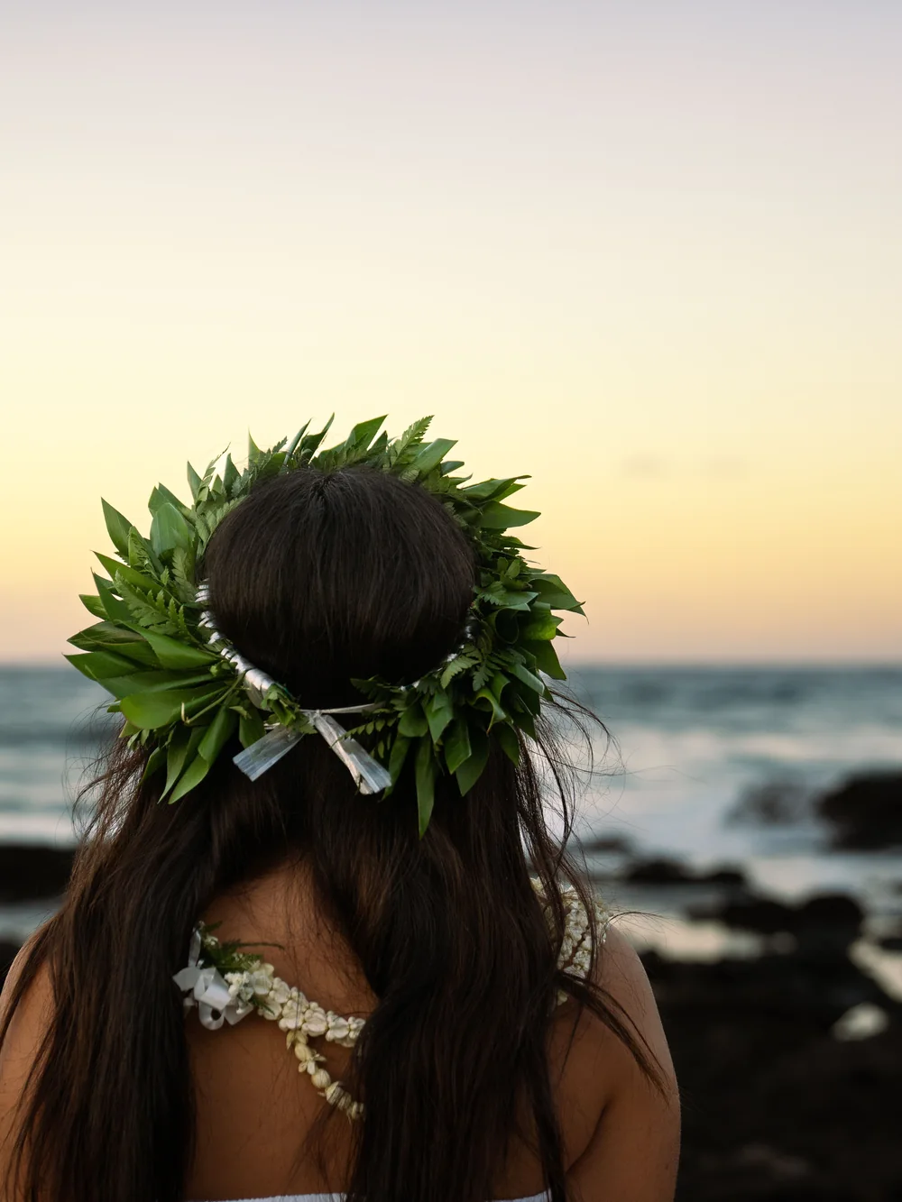 A woman wearing a leaf and flower lei looks out over the ocean at dusk, a dark headland in the distance